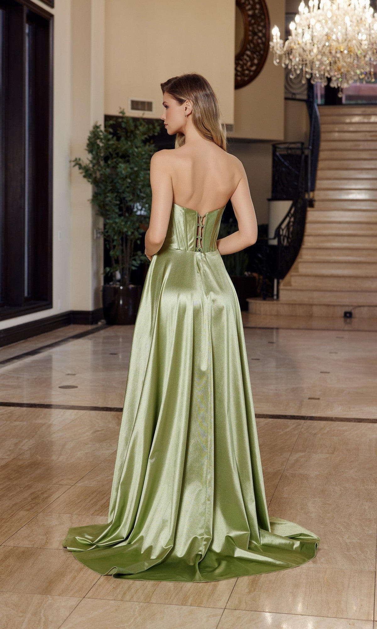 A woman stands indoors with her back to the camera, wearing the Nox Anabel K1735 long prom dress—a strapless satin gown with a corset bodice and elegant A-line skirt—in a marble-floored room.